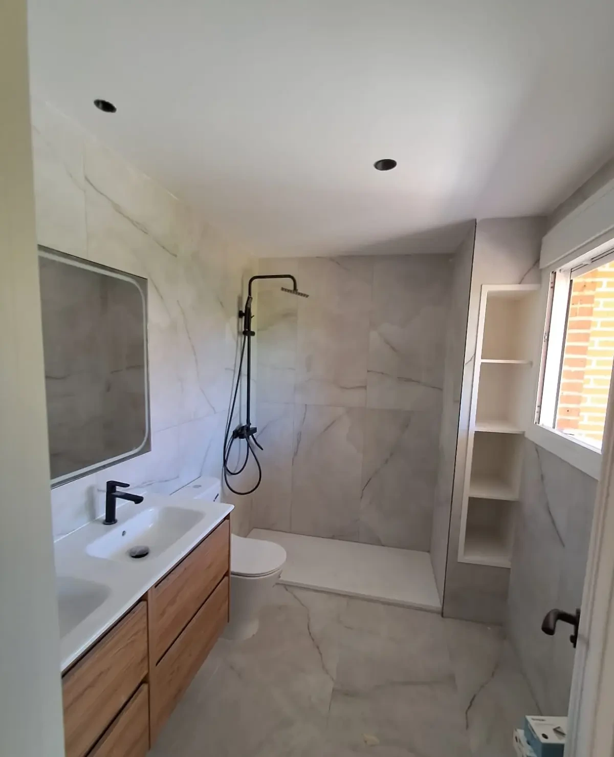 Reforma de baño con revestimiento de mármol blanco, mueble de madera y grifería negra en Madrid