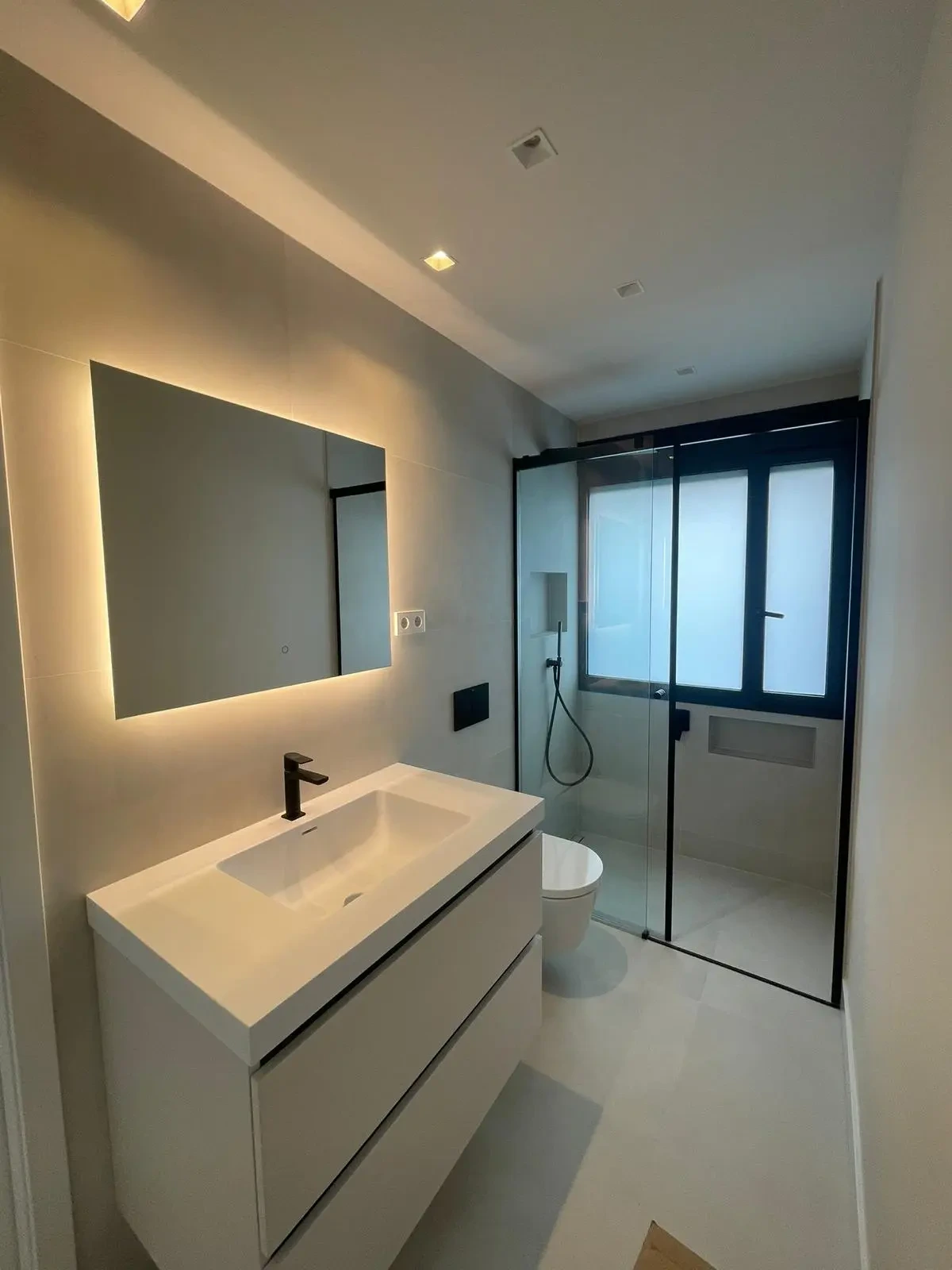 Reforma de baño con espejo retroiluminado, mueble blanco y mampara de perfilería negra en Madrid