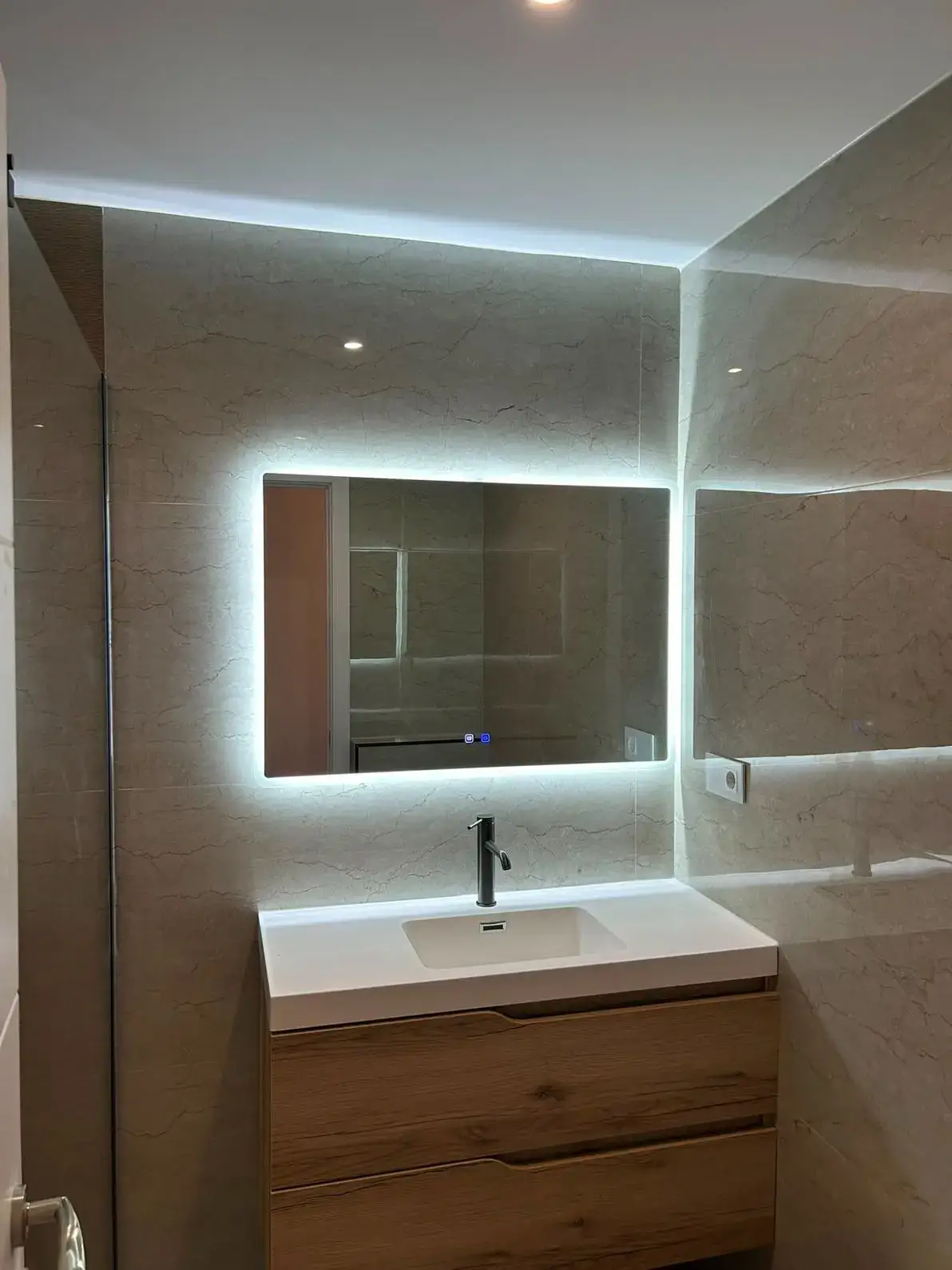 Reforma de baño con espejo retroiluminado sobre mueble de madera y paredes de mármol en Madrid