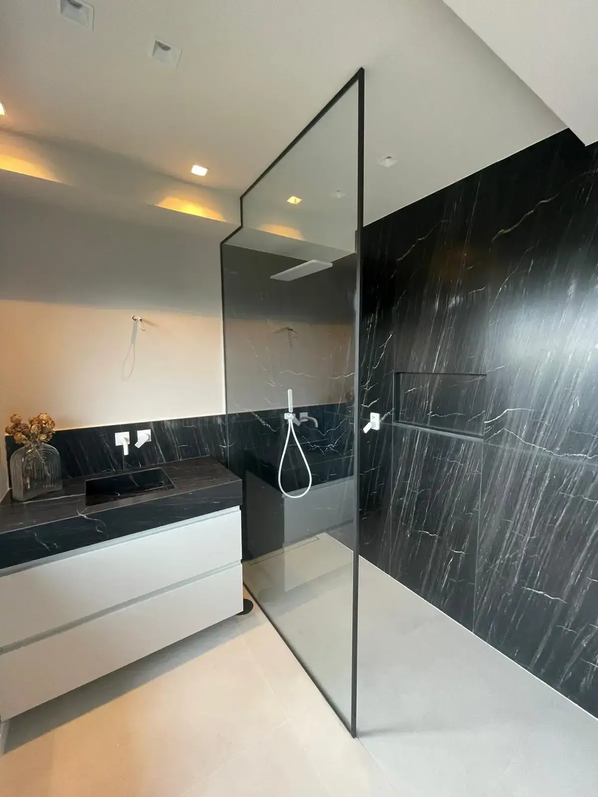 Reforma de baño con revestimiento de mármol negro, mampara perfilada y mueble blanco en Madrid